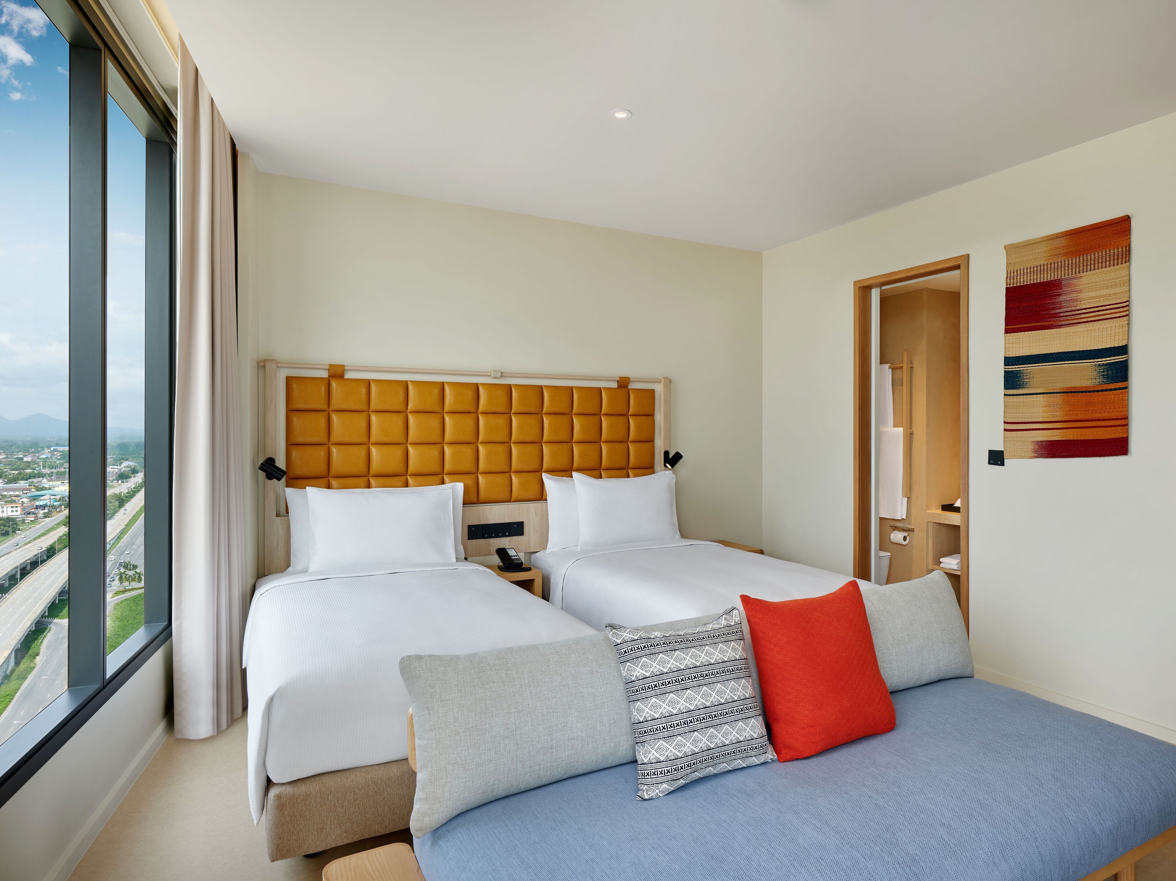A hotel room with two beds, a large window with a city view, a wooden headboard, and a wall-mounted artwork.