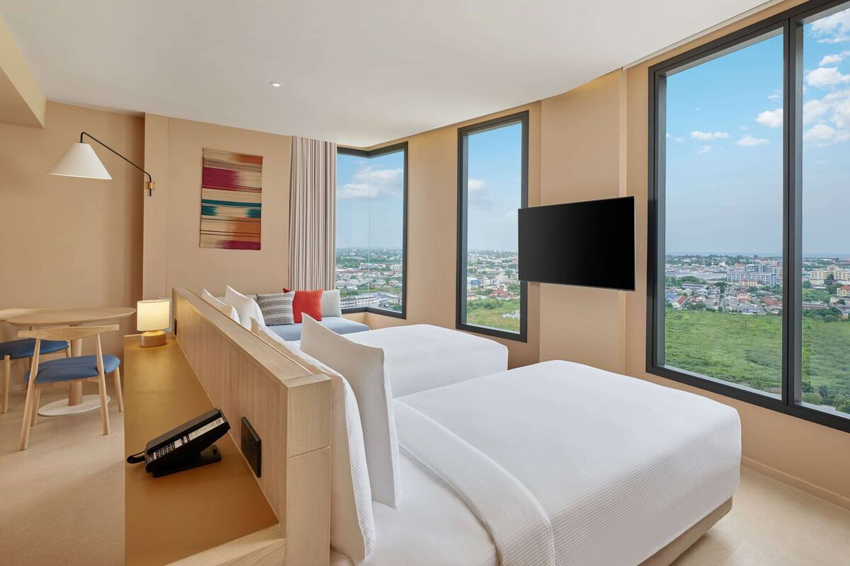 Habitación de hotel con dos camas, un escritorio, una silla, teléfono, televisión y ventanas amplias con vista a la ciudad.