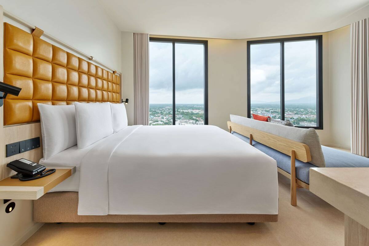 Una habitación de hotel moderna con una cama grande, un cabecero de madera, un escritorio con teléfono y vistas a la ciudad a través de amplios ventanales.