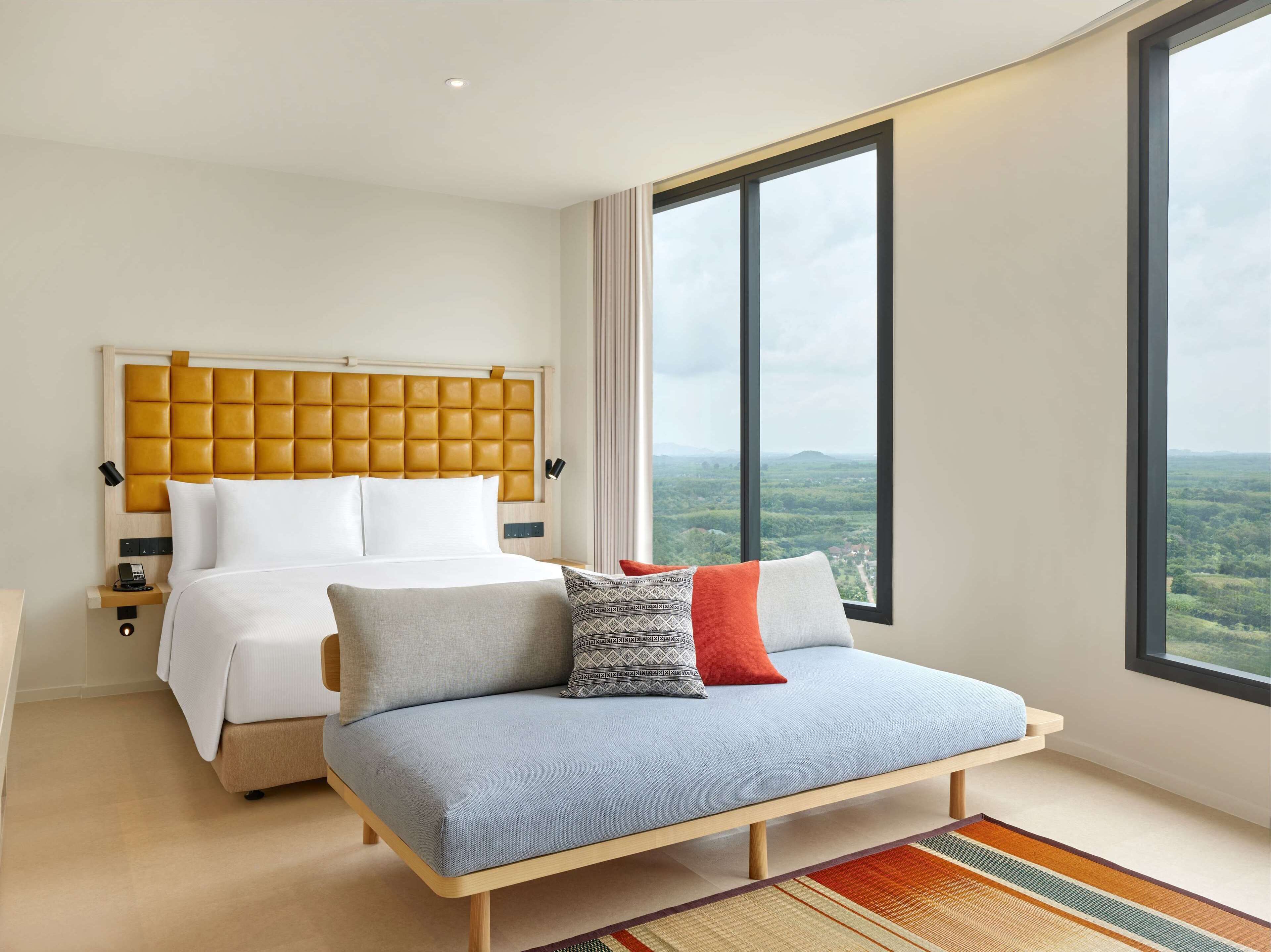 A modern bedroom with a large bed, a sofa bed, a wooden headboard, and a large window offering a view of the countryside.