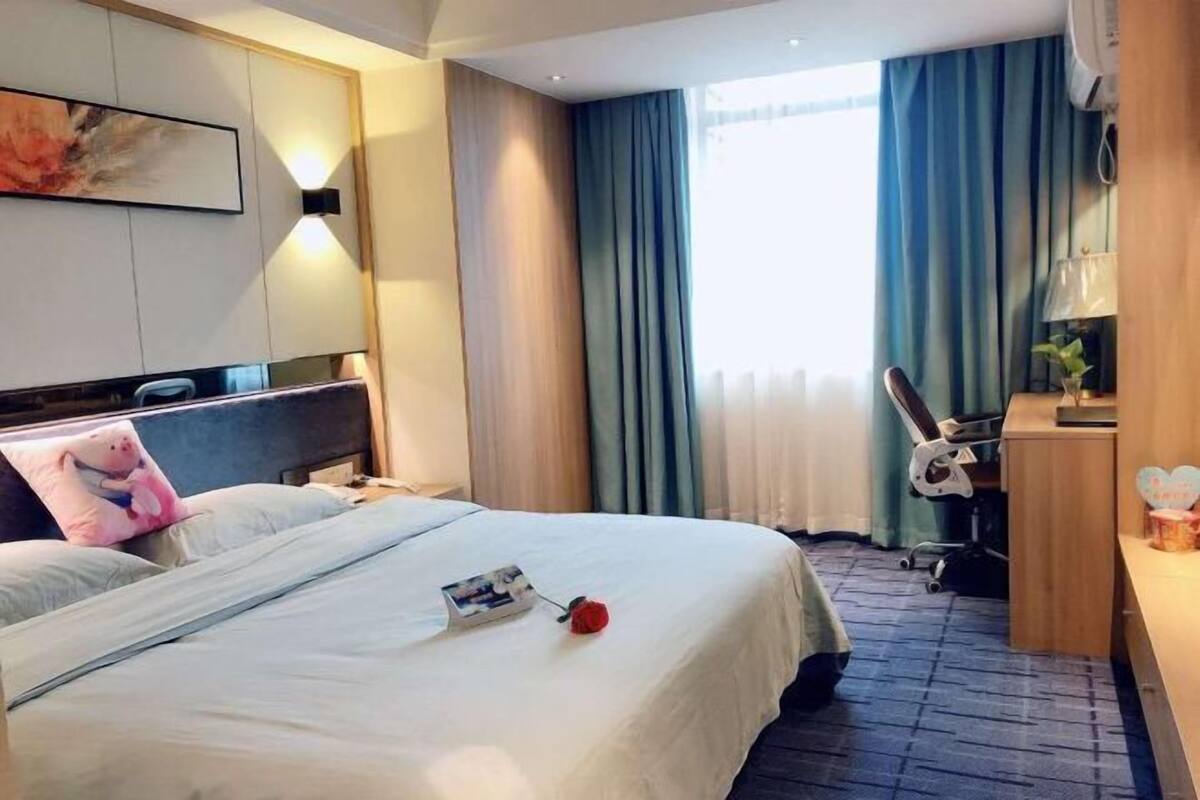 Una habitación de hotel con cama, escritorio, silla, lámpara y ventana con cortinas.