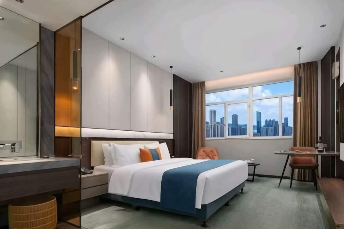 A hotel room with a bed, a desk, a chair, a window with a city view, and a bathroom.