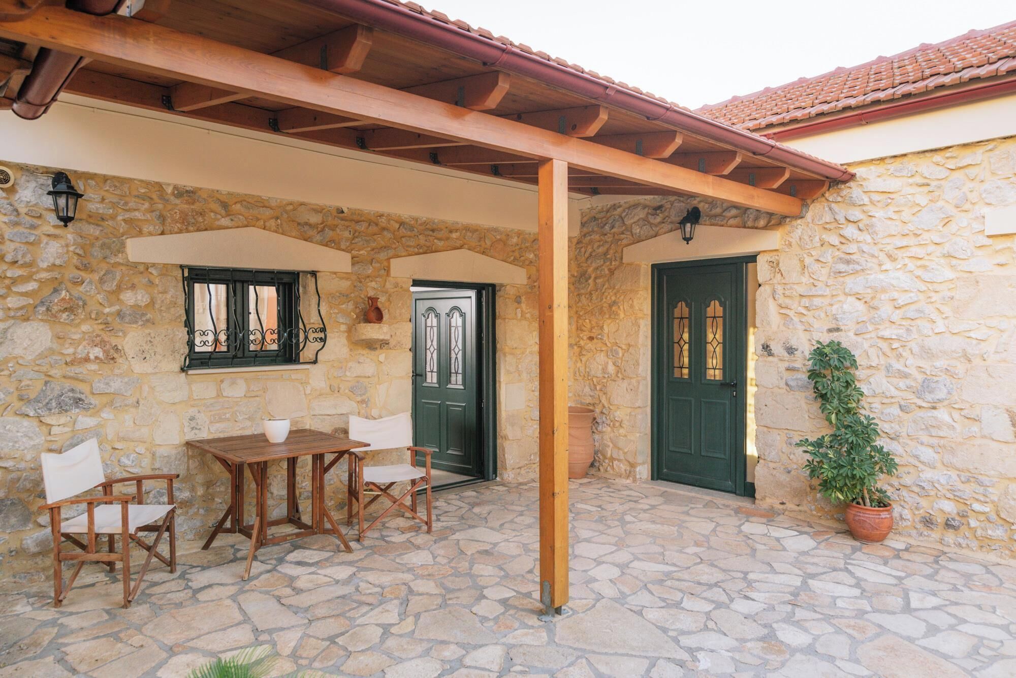 Ein Steinhaus mit einer Holzpergola, einem Tisch mit Stühlen, einer grünen Tür und einem Blumentopf.