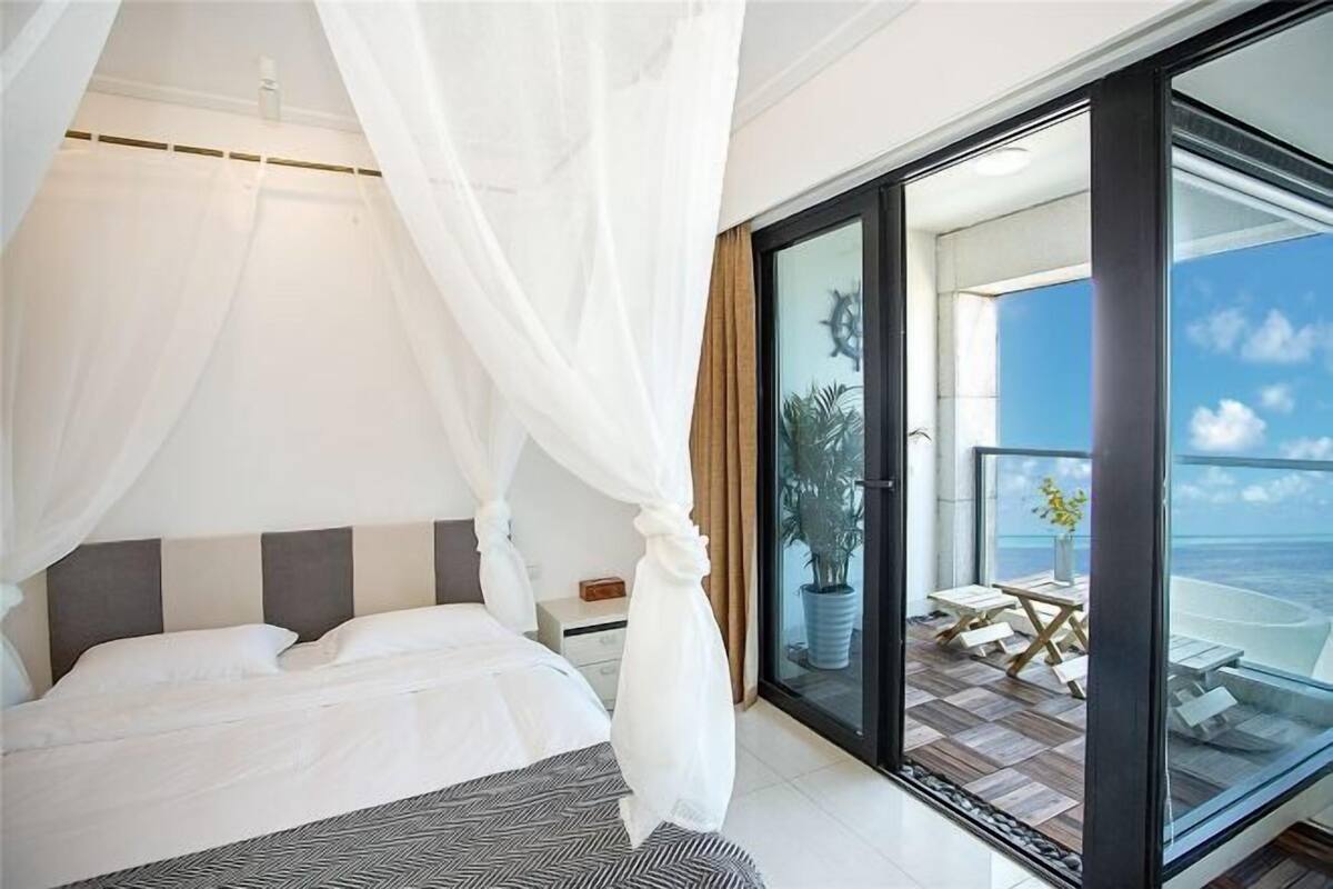 A bedroom with a bed, a nightstand, a bedside table, a large window, and a view of the outdoors.