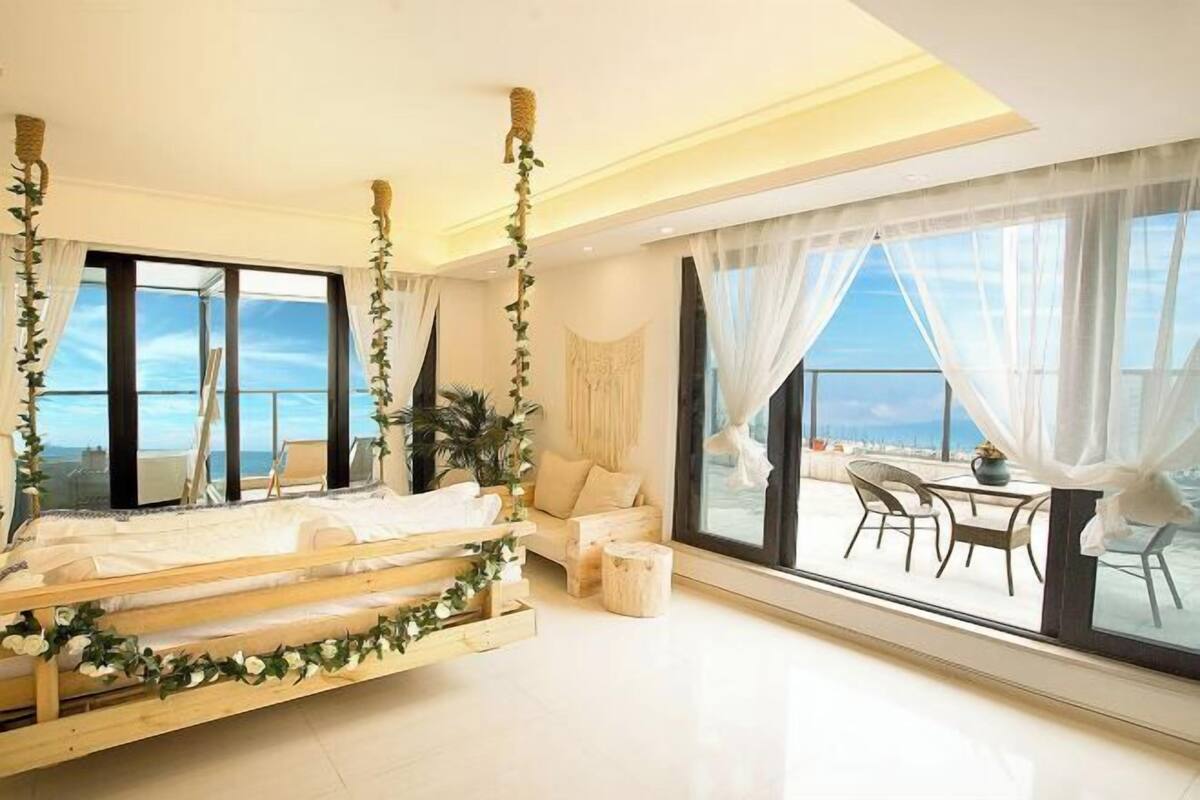 A modern bedroom with a large bed, a sofa, and a dining area with a view of the sea.