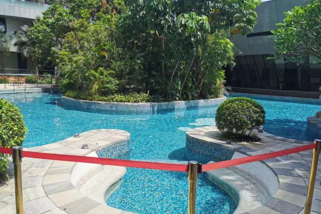 Una piscina con una barrera roja, rodeada de follaje y un edificio moderno al fondo.