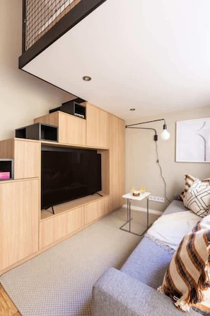 A modern living room with a large flat-screen TV, wooden cabinetry, a small side table, and a sofa with patterned cushions.