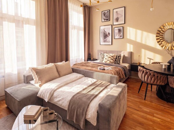 A modern bedroom with a large bed, a smaller bed, a desk, and a chair. There are curtains on the windows and a mirror on the wall.