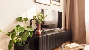 En moderne stue med et fladskærms-tv, en sort tv-reol, to potteplanter, et glasbord og et indrammet billede på væggen.