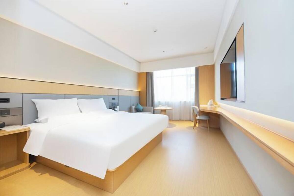 A modern hotel room with a large bed, bedside tables, a desk, and a large window.