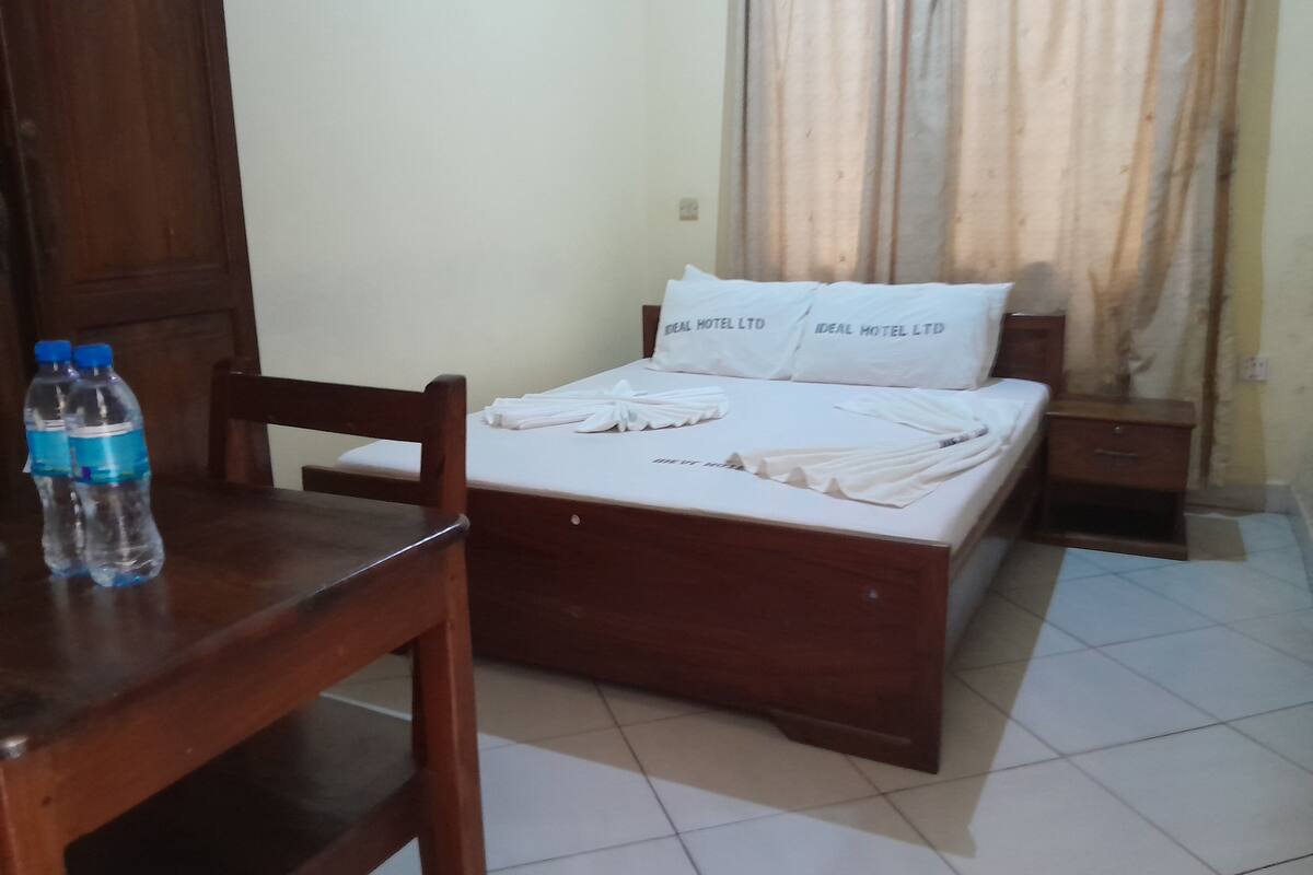 A hotel room with a wooden bed, a wooden nightstand, a wooden chair, and two water bottles on a wooden table.