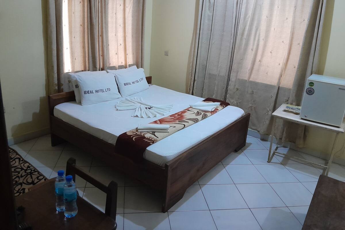 A neatly made bed with a floral bedspread and towels, a small table with water bottles, and a desk with a computer.