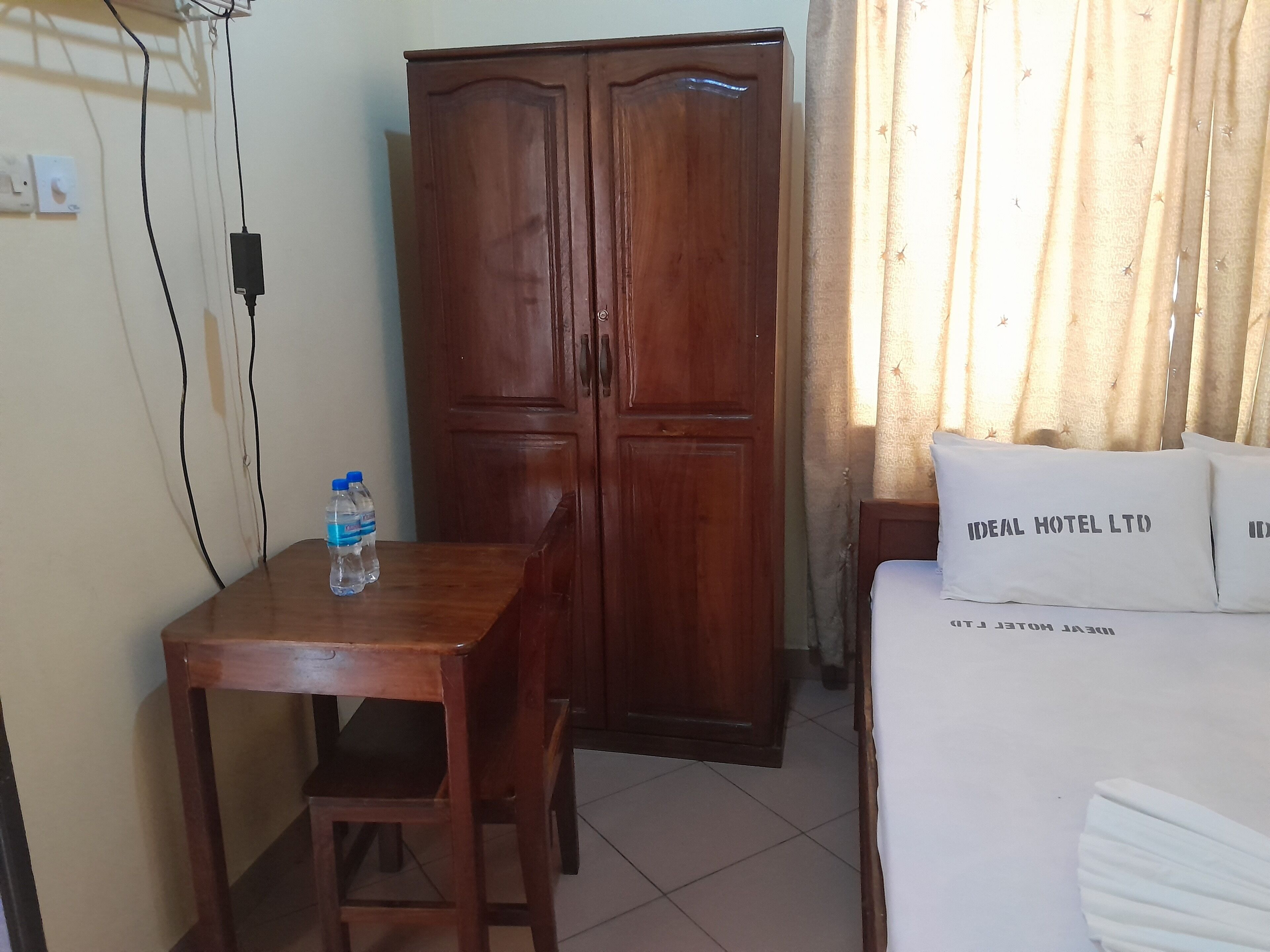 A hotel room with a wooden wardrobe, a wooden table with a water bottle, and a bed with pillows labeled "IDEAL HOTEL LTD".