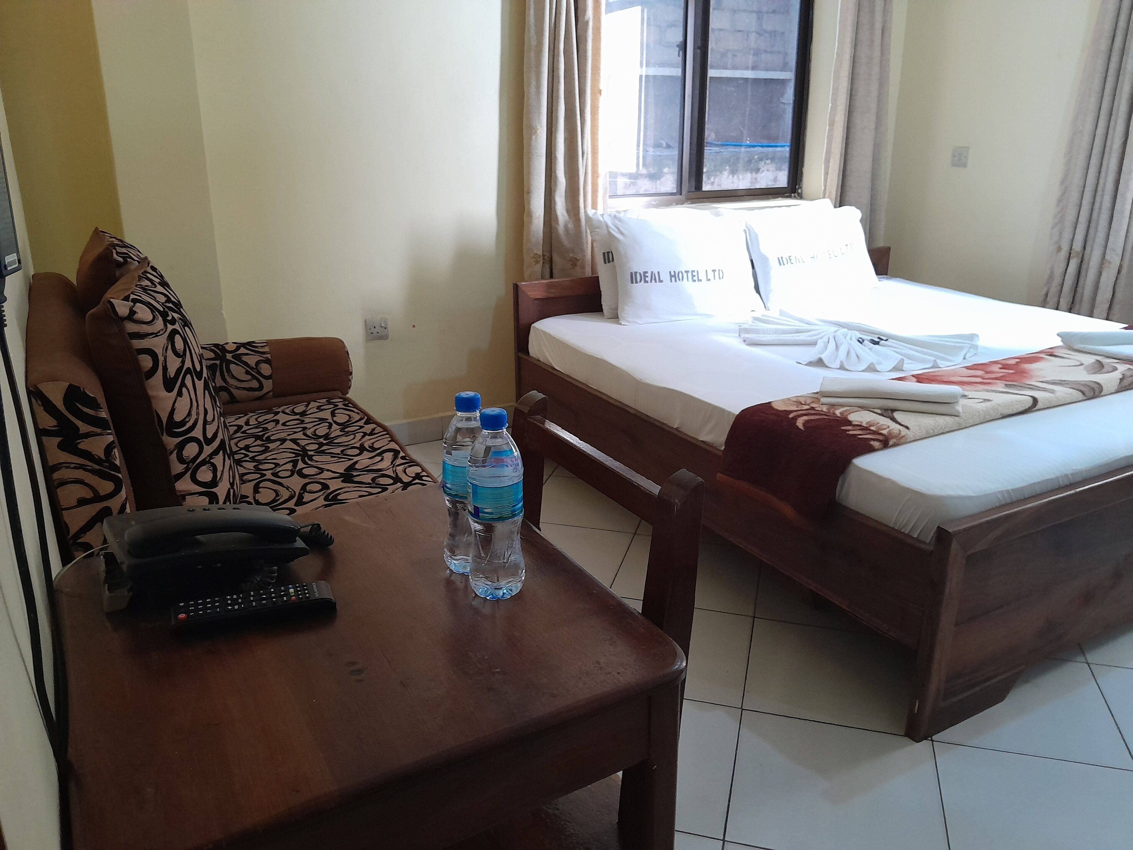 A hotel room with a bed, a sofa, a table with a phone and water bottles, and a window with curtains.
