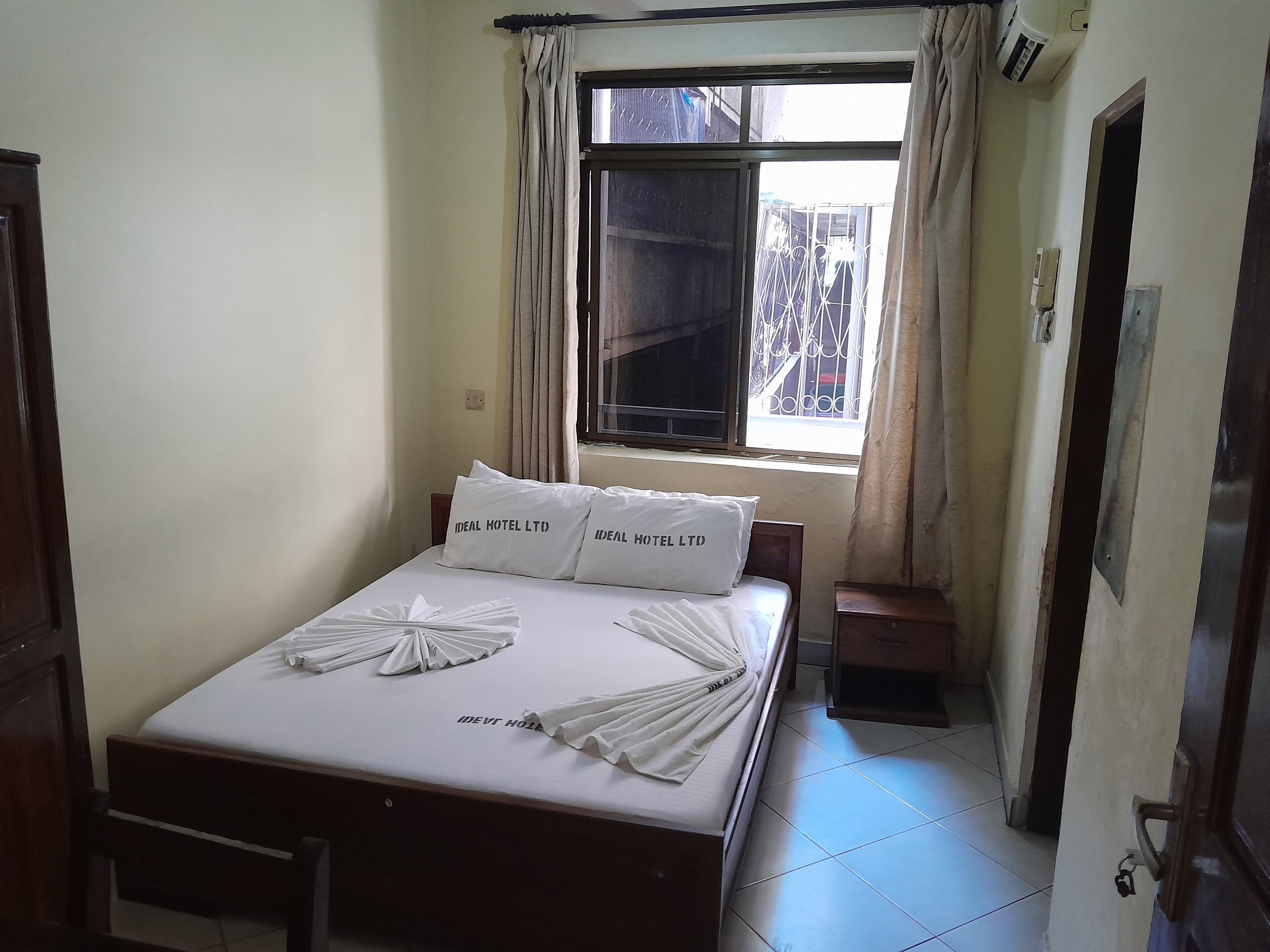 Cama bem arrumada com lençóis e almofadas brancas, janela com cortinas e uma pequena mesa de cabeceira.