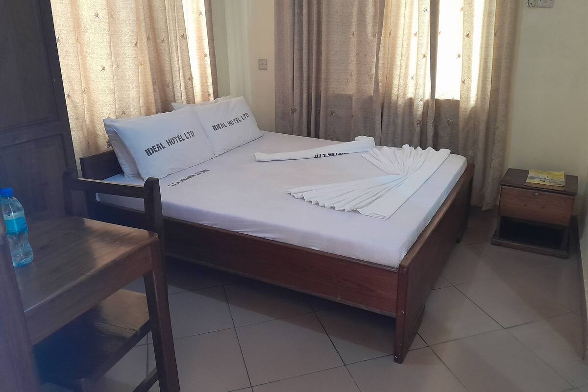 A neatly made bed with white linens and pillows, a wooden nightstand with a bottle and a small item on it, and a chair with a bottle beside it.