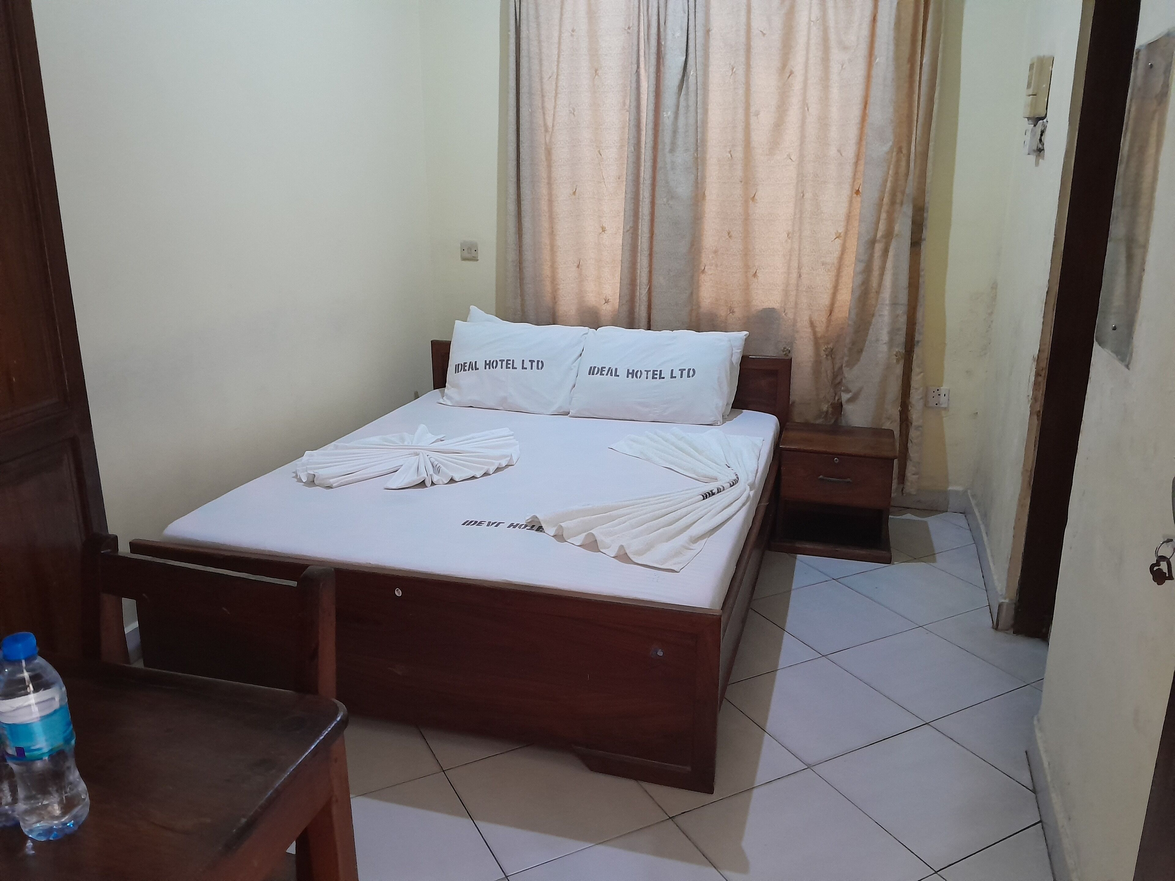 Cama bem arrumada com roupa de cama e toalhas brancas, mesa de cabeceira de madeira e uma garrafa de água em cima da mesa.