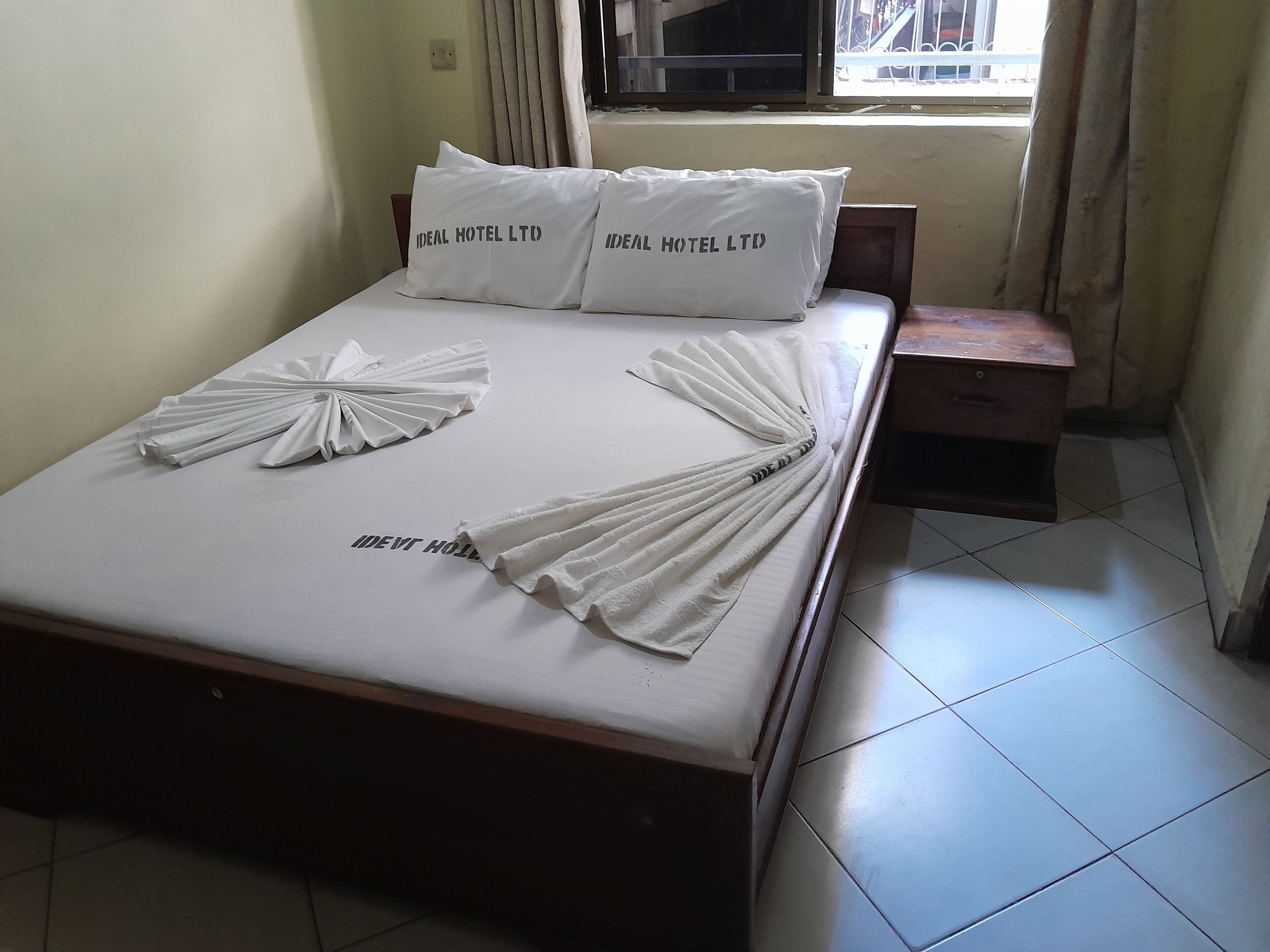 Un lit bien fait, avec du linge de lit blanc et des oreillers portant l’inscription « IDEAL HOTEL LTD. ». Deux serviettes décoratives sont disposées sur le lit.