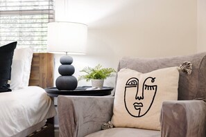 Un dormitorio con una cama, una silla con almohada para el cuello, una mesita de noche con lámpara y una ventana con persianas.