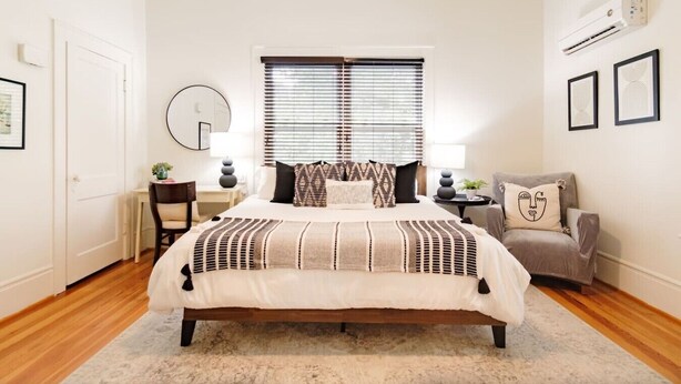 A bedroom with a bed, bedside tables, a chair, a window with blinds, and an air conditioning unit. - Black Velvet King Suite in Beautiful Highland Park (Birmingham)