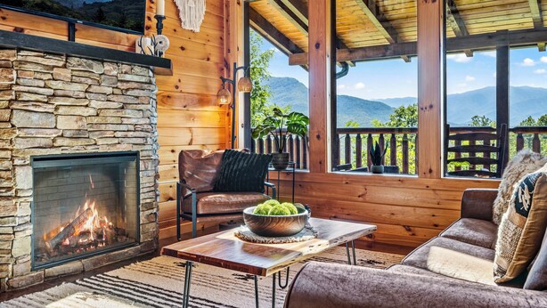 Un intérieur de chalet chaleureux avec une cheminée en pierre, des fauteuils en cuir et une vue sur les montagnes à travers de grandes fenêtres.