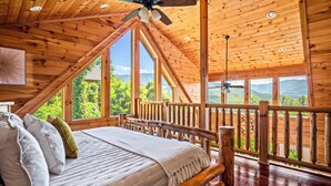 Une chambre située dans une cabane en bois, avec un lit, un ventilateur de plafond et de grandes fenêtres donnant sur les montagnes.