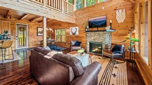 Une cabane en bois spacieuse avec une chambre à coucher en mezzanine, une cheminée en pierre et un grand salon doté de canapés en cuir et d’un téléviseur.
