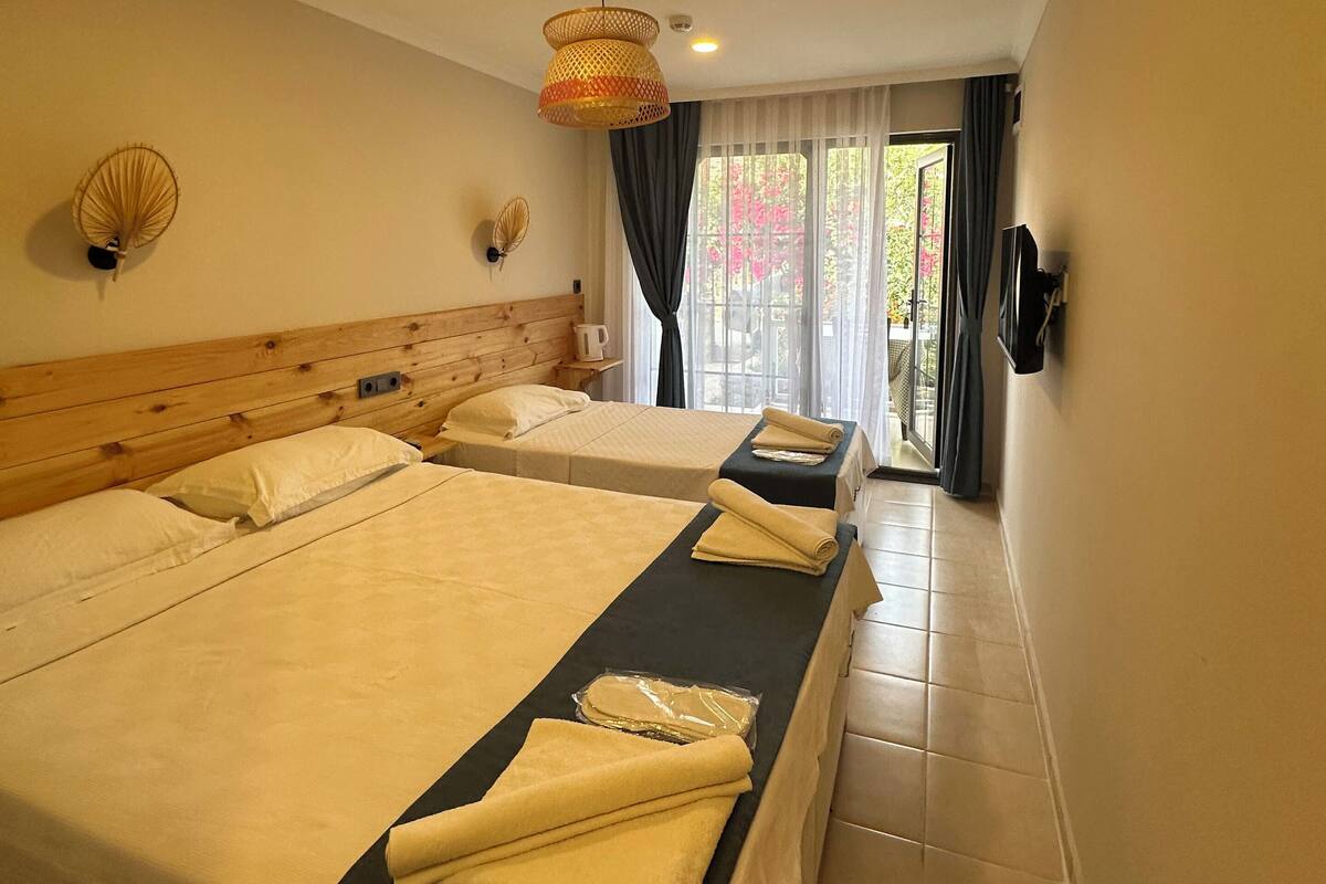 A hotel room with two beds, a wooden headboard, a ceiling fan, a wall-mounted TV, and a view of the outdoors through a window.