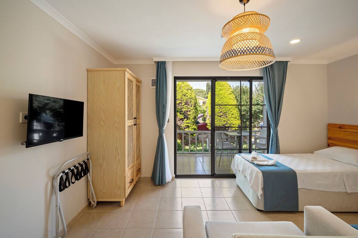 A hotel room with a bed, a TV, a wardrobe, a balcony with a view, and a hanging light fixture.