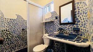 Un baño con paredes de mosaicos, un inodoro, un lavabo con espejo y una ventana sobre el lavabo.