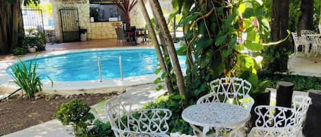 Zona de piscina con sillas y mesas de hierro forjado blanco, rodeada de exuberante follaje y una estructura con techo de paja.