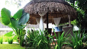 Un área de estar al aire libre con techo de paja, un banco de madera y una mesa, rodeada de exuberante follaje y hojas grandes.