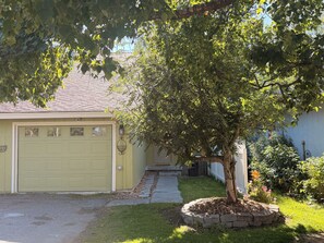 A residential garage with a tree in front, a pathway leading to the garage, and a grassy yard with a flower bed. - Entire Home: Westchester Lagoon (Anchorage)