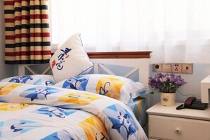 Un lit bien fait, avec une literie à motifs, une table de chevet sur laquelle se trouvent un téléphone et un pot de fleurs, et un rideau à rayures.