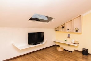 A modern living room with a flat-screen TV on a white shelf, a skylight, a small round ottoman, and a trash can.