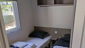 Ein kleines, schlichtes Zimmer mit zwei Einzelbetten, einem Fenster und einem Regal über den Betten.