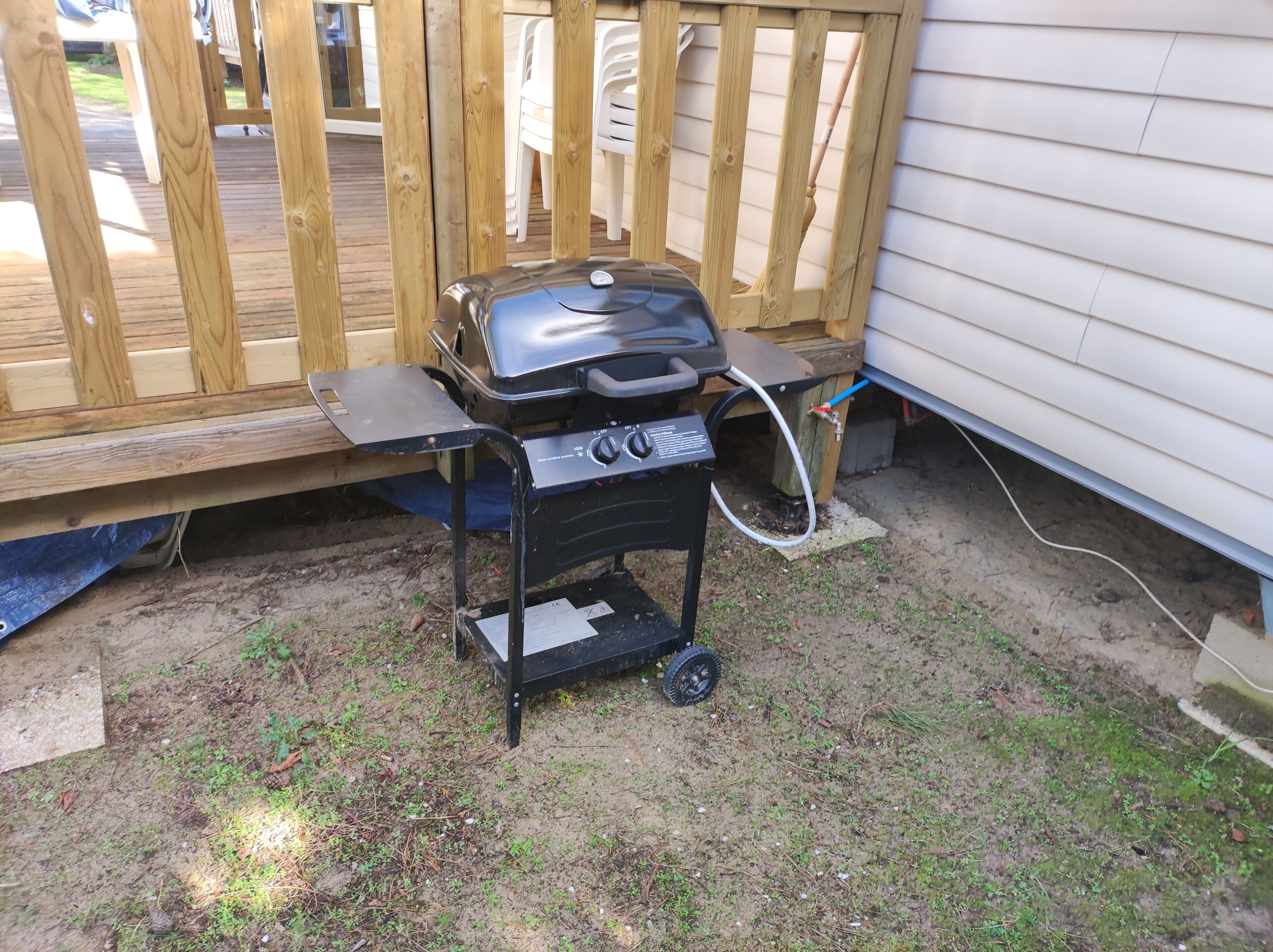 Una parrilla de barbacoa portátil con ruedas, ubicada sobre terreno de tierra al lado de una terraza de madera y una casa.