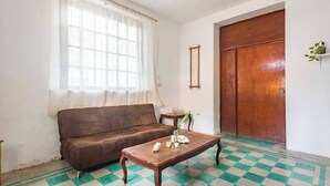 Une pièce avec un canapé marron, une table basse en bois agrémentée d’objets décoratifs, une grande fenêtre avec des rideaux légers, une porte en bois et un sol à carreaux.