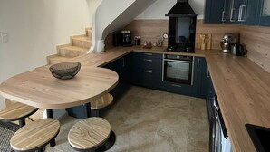 A modern kitchen with a wooden dining table and stools, a built-in oven, and dark cabinetry. - Maison à la Hauteur d un Beau Village (Apt)