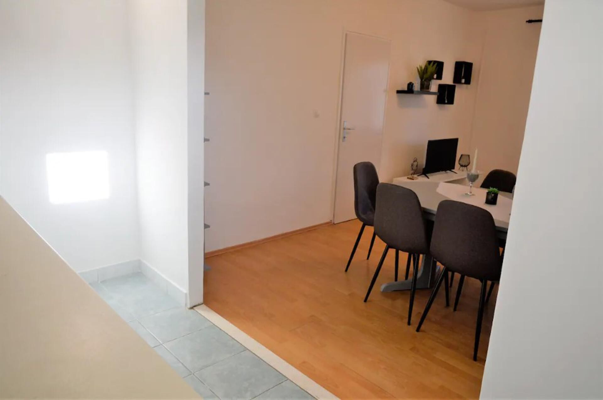 A dining area with a table and four chairs, a white door, and a small shelf with decorative items.