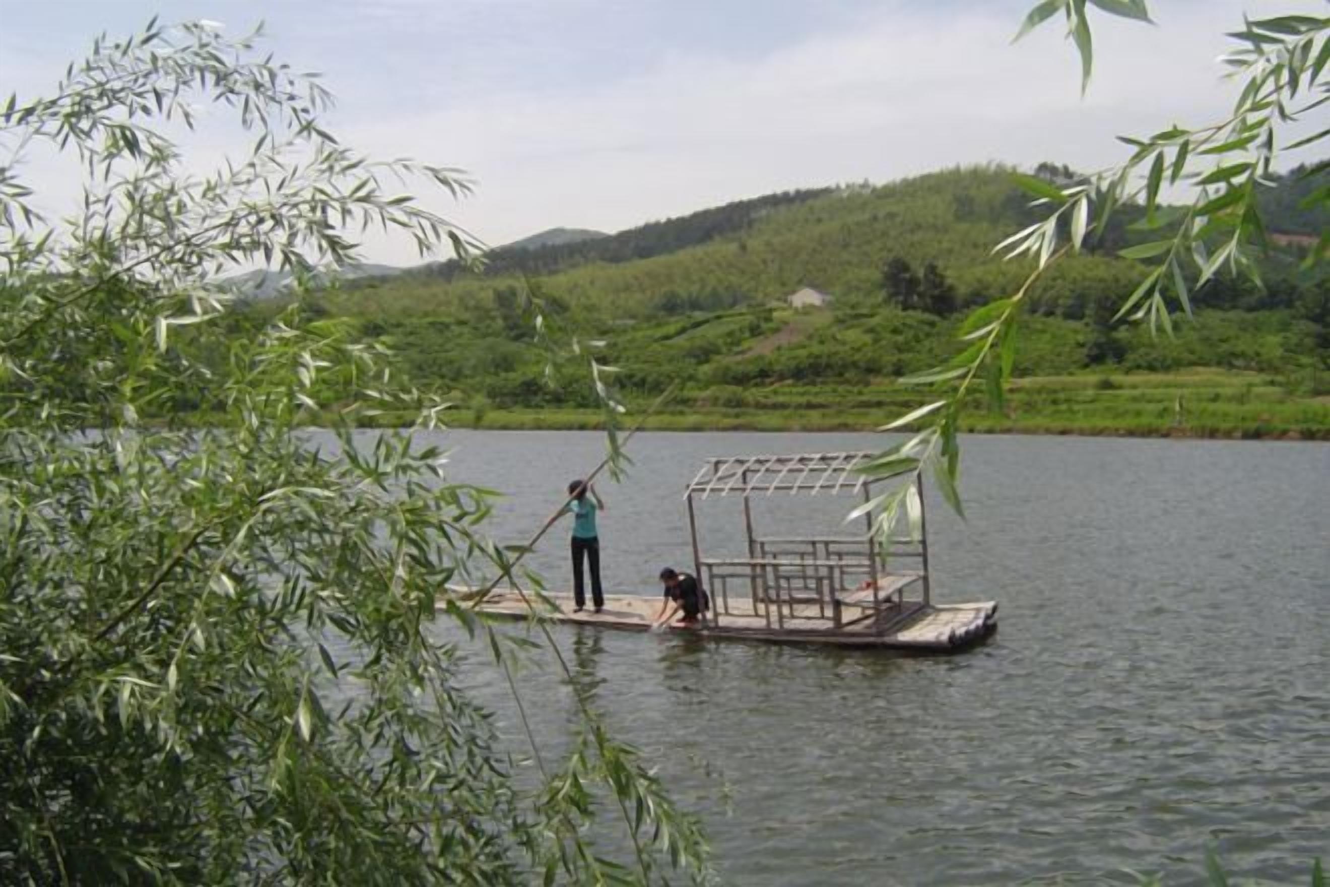 Un radeau en bambou sur un lac paisible, avec deux personnes à bord, entouré d’une végétation luxuriante et avec des collines en arrière-plan.