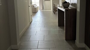 A hallway with a patterned rug, a dining area with a bowl of fruit, and a wall with a geometric art piece.