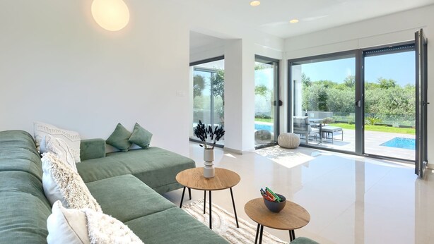 Una sala de estar moderna con un sofá verde, dos mesitas de madera y un amplio ventanal que da a un patio exterior con piscina.