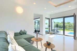A modern living room with a green sofa, two wooden side tables, and a large glass door leading to an outdoor patio with a pool. - Luxury villa with pool, barbecue area 1 km to the beach (Liznjan)