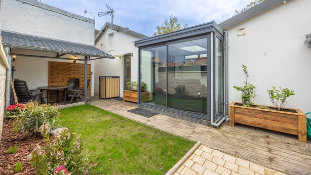 Ein moderner Patio-Bereich mit Glasveranda, einem Holztisch und -stühlen, sowie ein kleiner Garten mit Pflanzen und Blumen.