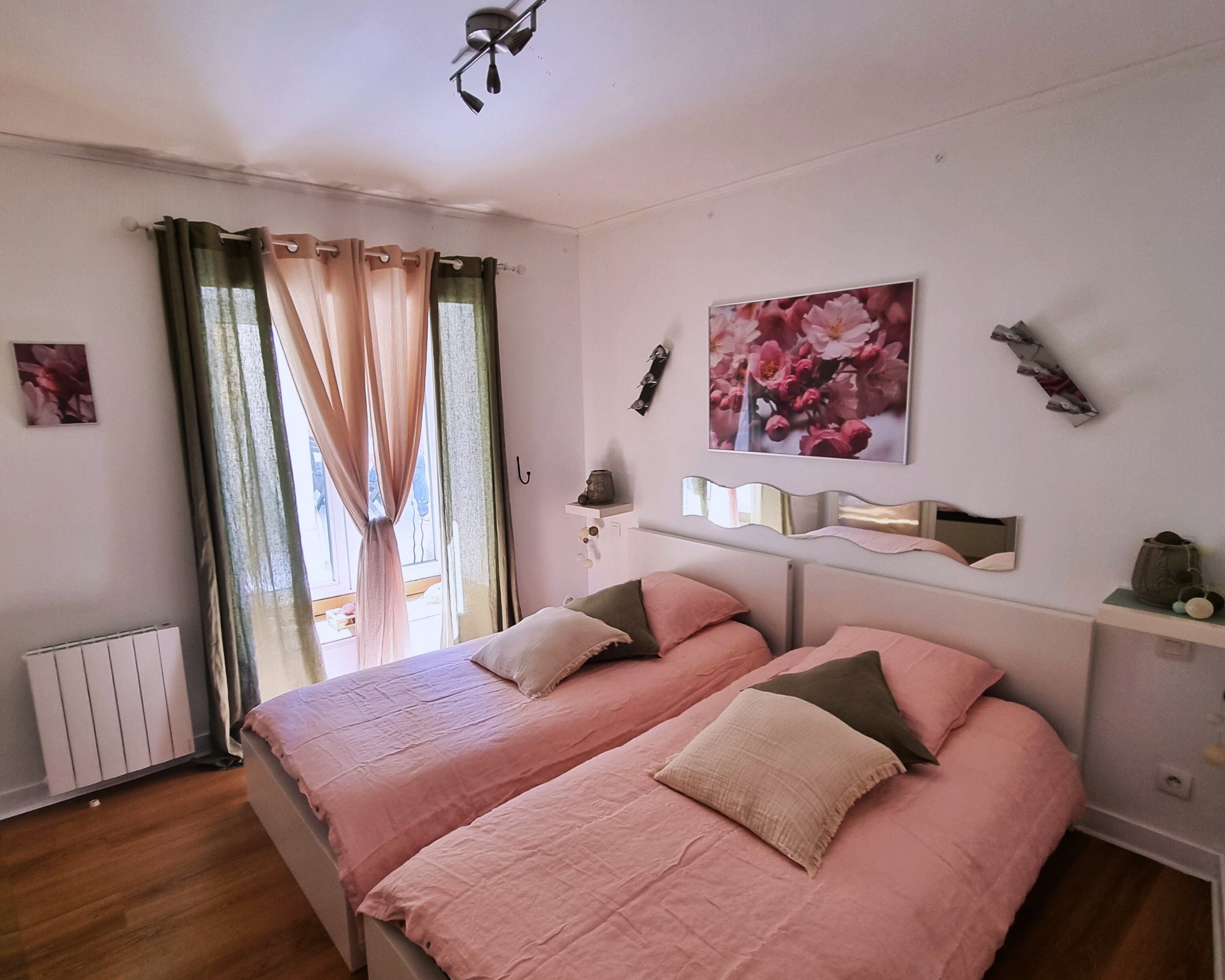 A bedroom with two beds, a window with curtains, a radiator, and a wall-mounted shelf with decorative items.