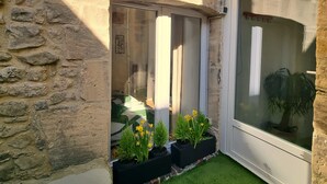 Un bâtiment en pierre avec un petit jardin, une fenêtre donnant sur un arbre et une porte dotée d’un cadre blanc.
