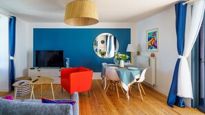 Una sala de estar moderna con una pared azul de acento, un espejo redondo, una mesa de comedor con sillas, un sillón rojo y un televisor sobre un mueble blanco.