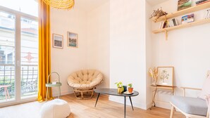 A bright living room with a large window, a round white ottoman, a beige armchair, a small black table with plants, and a bookshelf with books and decorative items. - L'Atypique - Appt à 150m de la plage (Arcachon)