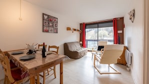A cozy living room with a dining table, chairs, a sofa, and a window with red curtains. - Le Corbier - Studio avec belle vue montagne (Villarembert)